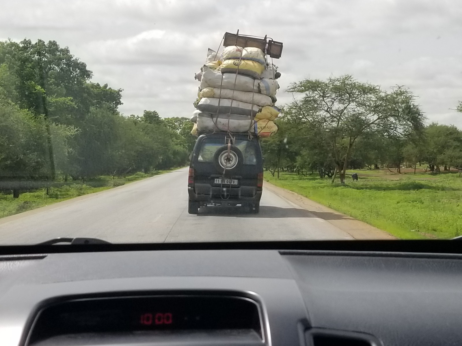A van on a rural road loaded high with sacks of cargo, seen through a car windshield in Burkina Faso